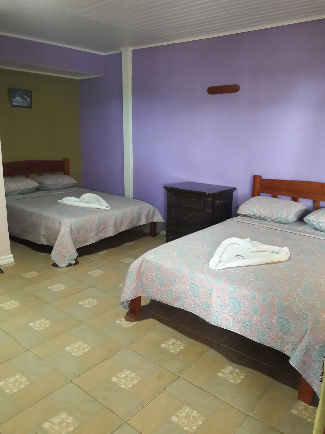A room with two beds, each with a floral-patterned bedspread and a towel on the bed. There is a dark-colored dresser and a light-colored wall.