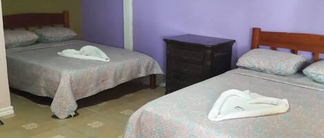 A room with two beds, each with a floral-patterned bedspread and a towel on the bed. There is a dark-colored dresser and a light-colored wall.