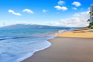 Une plage à l’eau d’un bleu limpide, bordée d’un rivage sablonneux, avec des montagnes qui se dessinent à l’horizon.