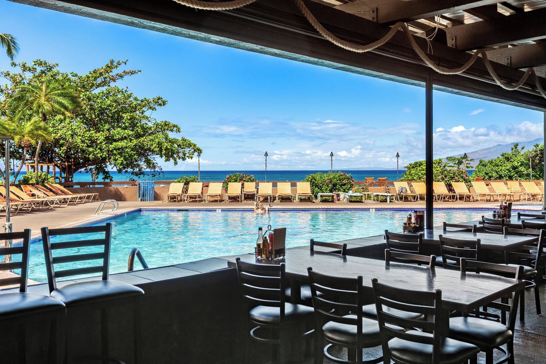 Un espace aménagé au bord de la piscine, avec des chaises longues et des tables, offrant une vue sur un océan d’un bleu limpide.