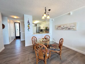 A dining area with a round glass table and four wicker chairs, a kitchen with white cabinets, and a hallway leading to a door with frosted glass.