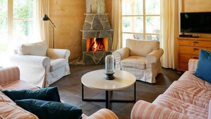 Un salon chaleureux avec une cheminée en pierre, deux fauteuils blancs, un canapé à rayures, une table basse ronde et un téléviseur posé sur un meuble en bois.