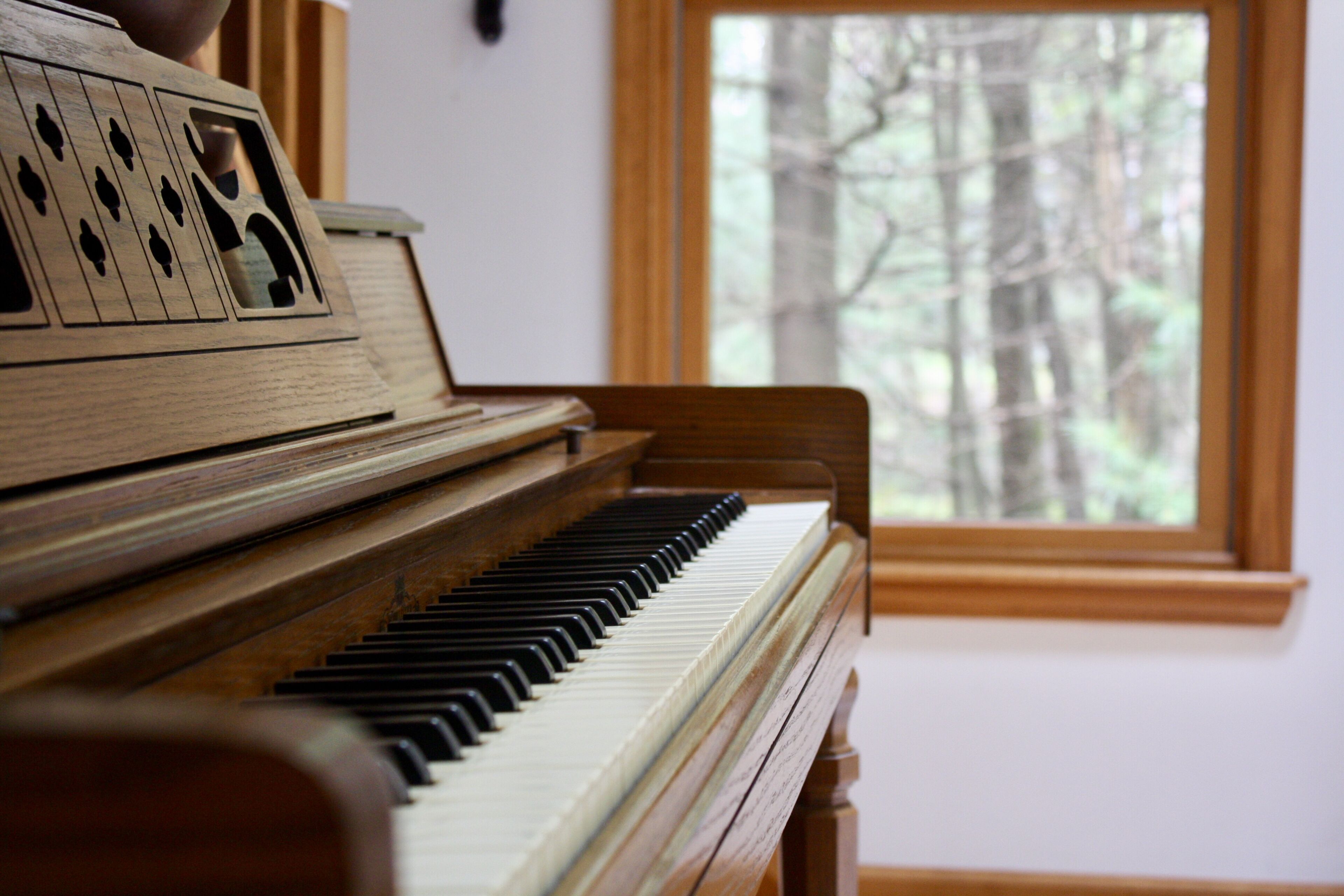 A wooden piano with a view of a forest through a window.