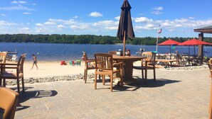 A beachside patio with wooden tables and chairs, a covered area with red umbrellas, and a view of a lake with boats. - ⭐Central heat/AC ⭐ WiFi ⭐ Near pool, lake, beach!⭐ (Pocono Pines)
