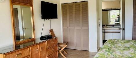 Un dormitorio con cómoda de madera, televisor de pantalla plana en la pared, cama con colcha floral y baño con lavamanos y espejo.