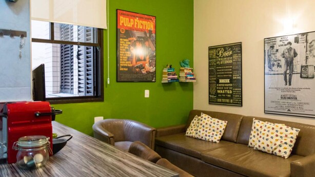A room with a green wall, a brown leather couch with patterned pillows, a wooden table with a red appliance, and posters on the wall. - LIVING ROOM HALF A BLOCK AWAY – CHANNEL 6 – AIR CONDITIONING, WI-FI, CABLE TV, WASHER/DRYER AND GARAGE (Santos)