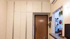 A room with a wooden door, white cabinets, a small table with a television, and a wall-mounted shelf with various items. - LIVING ROOM HALF A BLOCK AWAY – CHANNEL 6 – AIR CONDITIONING, WI-FI, CABLE TV, WASHER/DRYER AND GARAGE (Santos)