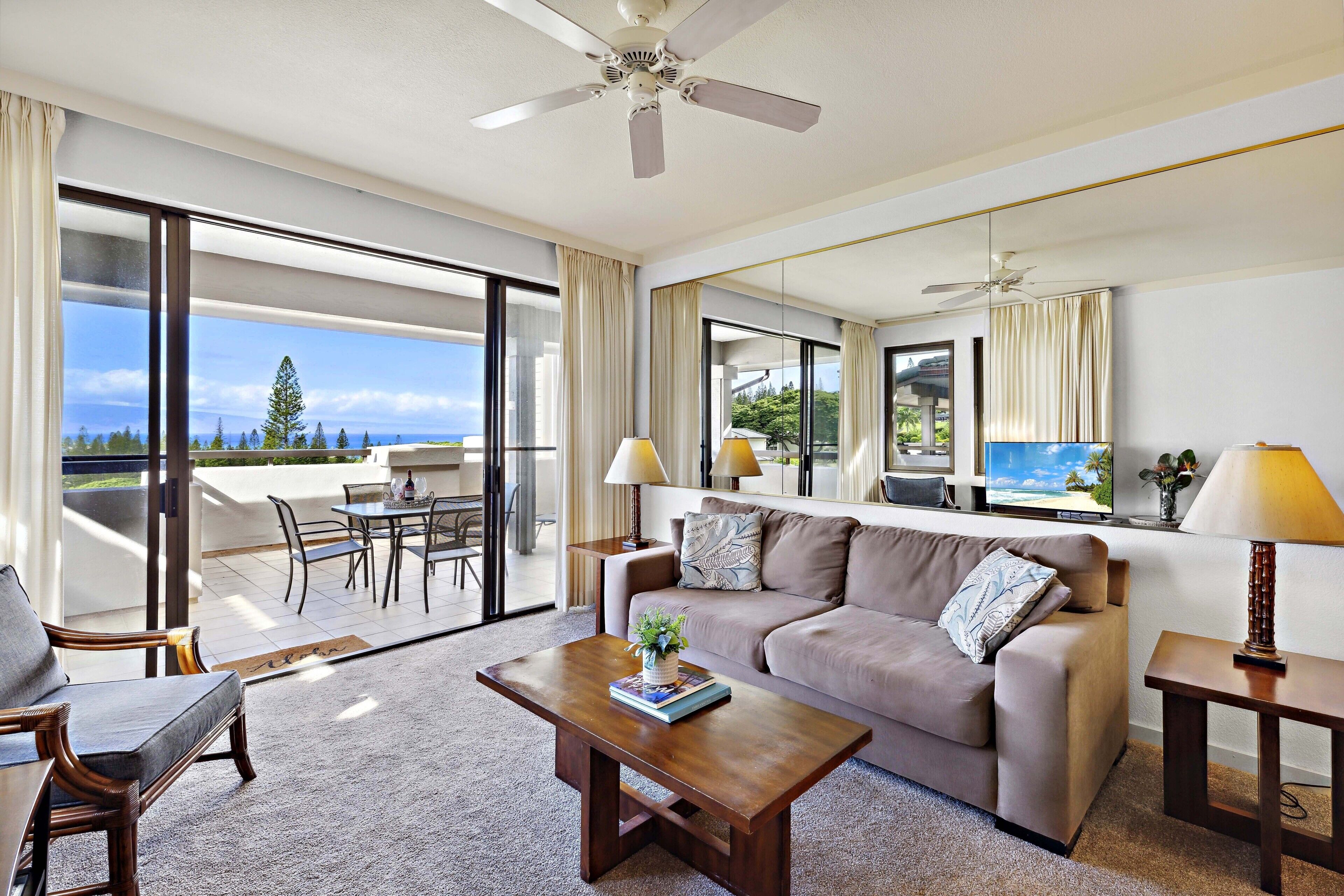 A spacious living room with a large sofa, a coffee table, and a television. There is a balcony with a table and chairs, and a view of the outdoors.