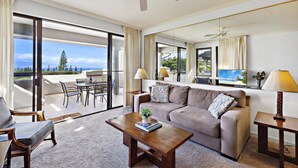 A spacious living room with a large sofa, a coffee table, and a television. There is a balcony with a table and chairs, and a view of the outdoors. - Great Deal $175/n (8/9-11/20) Ocean View AC-Kapalua Golf Villa 19T4-Sullivan (Lahaina)