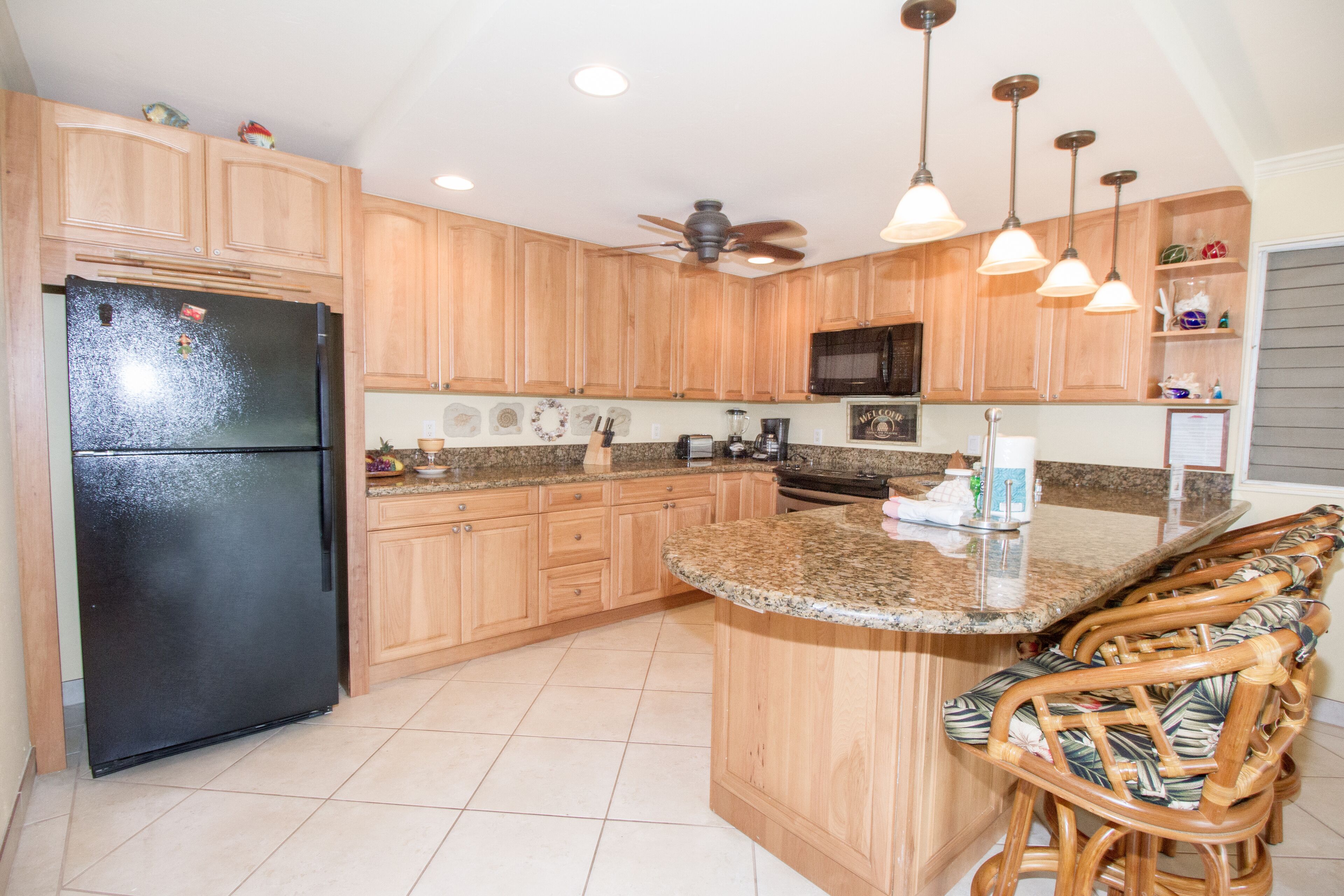 A kitchen with wooden cabinets, a black refrigerator, a microwave, a ceiling fan, and a bar area with chairs.