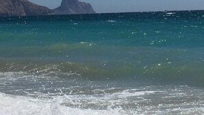 A beach with clear turquoise water, pebbles, and distant rocky cliffs.