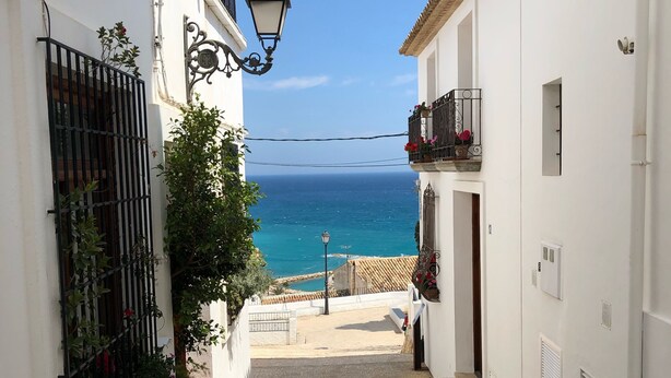 A narrow street leading to a coastal view with white buildings, a metal gate, and a street lamp.