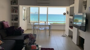 A living room with a view of the ocean, a sofa, a coffee table with a vase, and a TV on the wall.