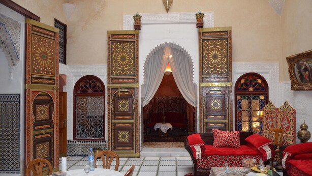 A traditional Moroccan interior with ornate doors, a sofa with red cushions, and a dining area with a table and chairs. - Traditional riad with modern comfort 6/8 per in the heart of the medina. (Meknès)