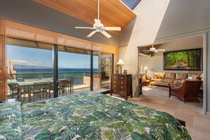 A spacious bedroom with a large bed, a balcony with a view of the ocean, and a living area with a sofa and a painting. - 3BD/3BA 5 Star! Beach & Sunsets Oceanview-SK474- Sullivan Properties (Lahaina)