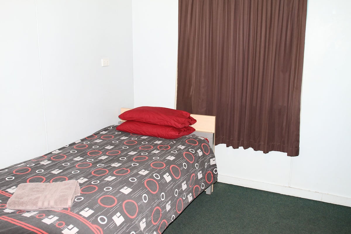 A small, single-bed room with a patterned bedspread, red pillows, and a brown curtain.