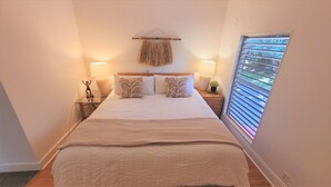 A bedroom with a large bed, two bedside tables, a hanging decor piece, and a window with blinds. - Spectacular Sunsets & Ocean Views with AC! $250/n 9/1-9/30  KS D2-Sullivan (Lahaina)
