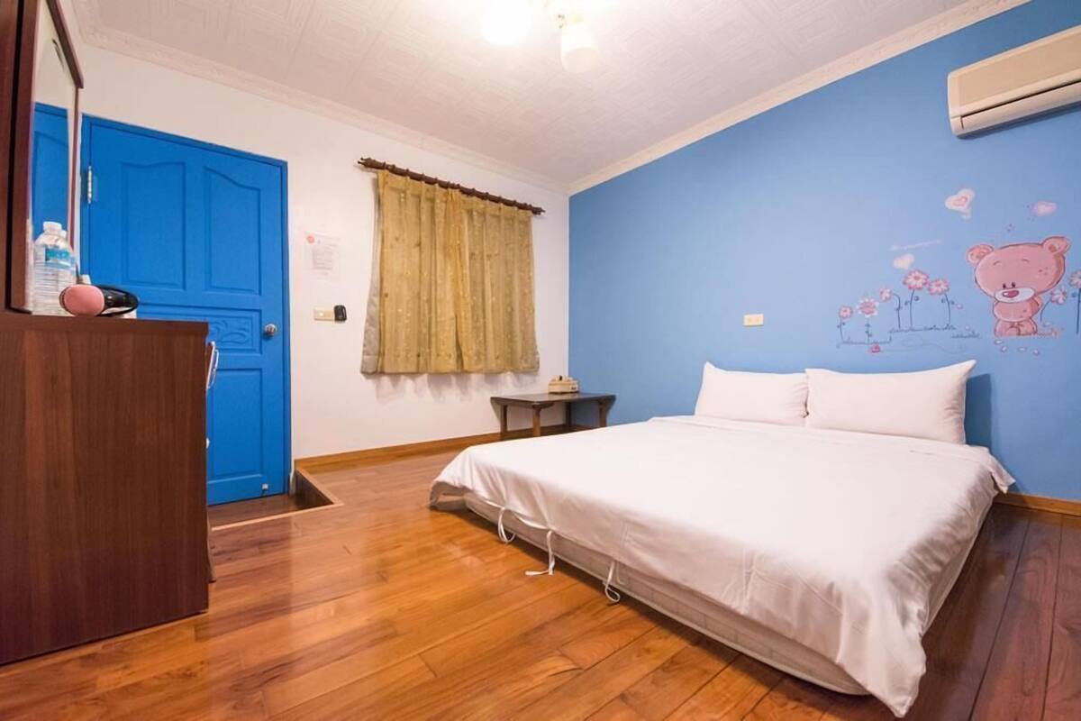 A bedroom with a wooden floor, a bed with white bedding, a blue wall with a bear mural, a blue door, and a window with curtains.