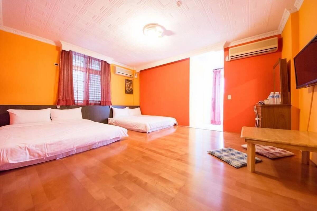A hotel room with two beds, a wooden floor, an air conditioning unit, a television, and a bench.