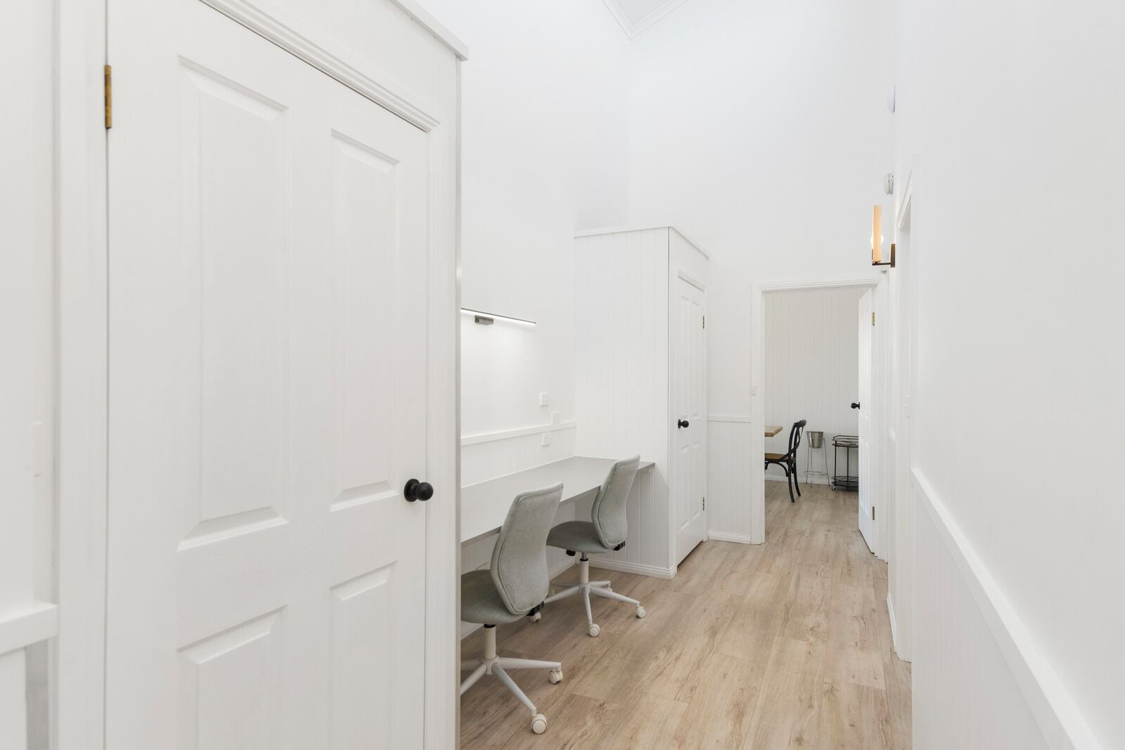 A hallway with white walls and doors, a desk with a chair, and a small table with chairs in the background.