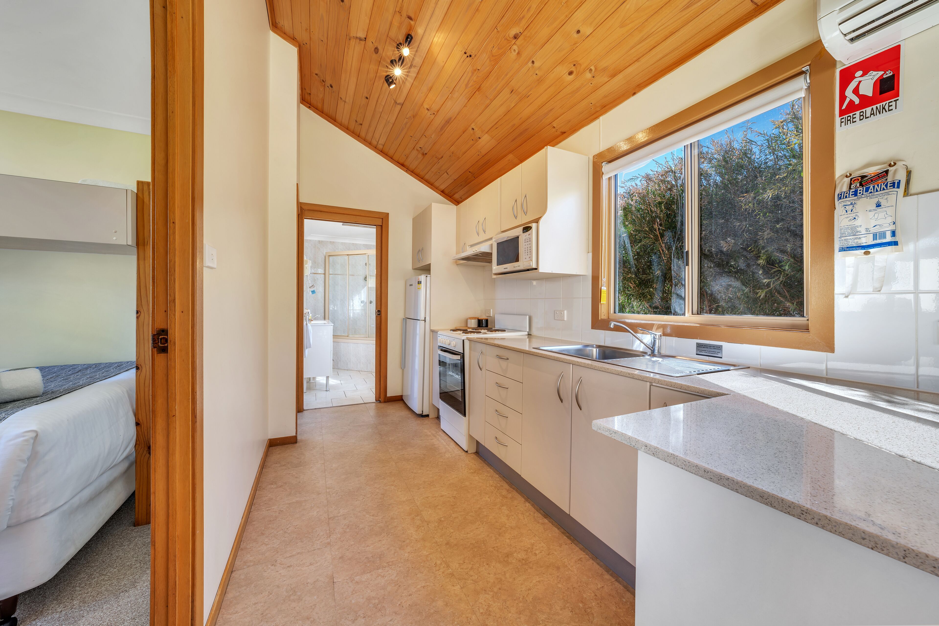 Cocina moderna con armarios blancos, microondas y lavamanos. Hay una ventana con vista a árboles y un cartel de manta ignífuga en la pared.