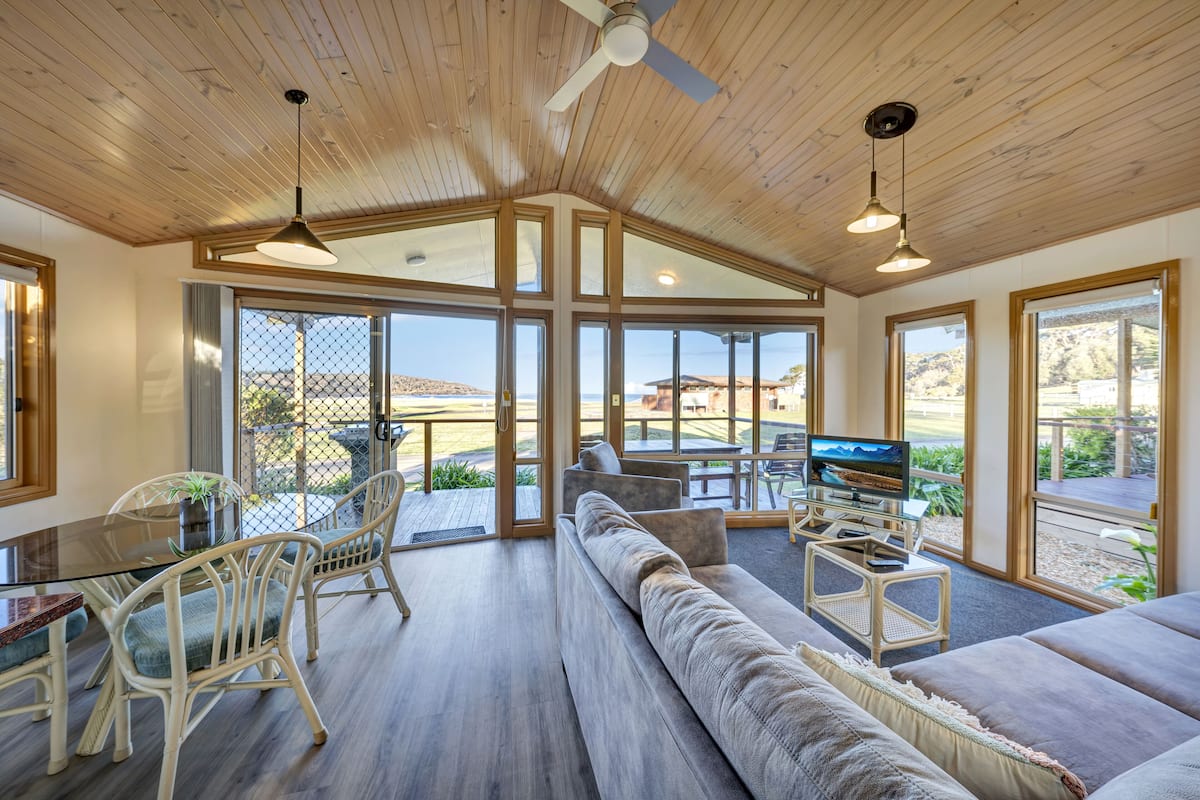 Amplia sala de estar con un sofá grande, mesa de comedor y sillas, televisor y ventanas grandes que ofrecen vistas al exterior.
