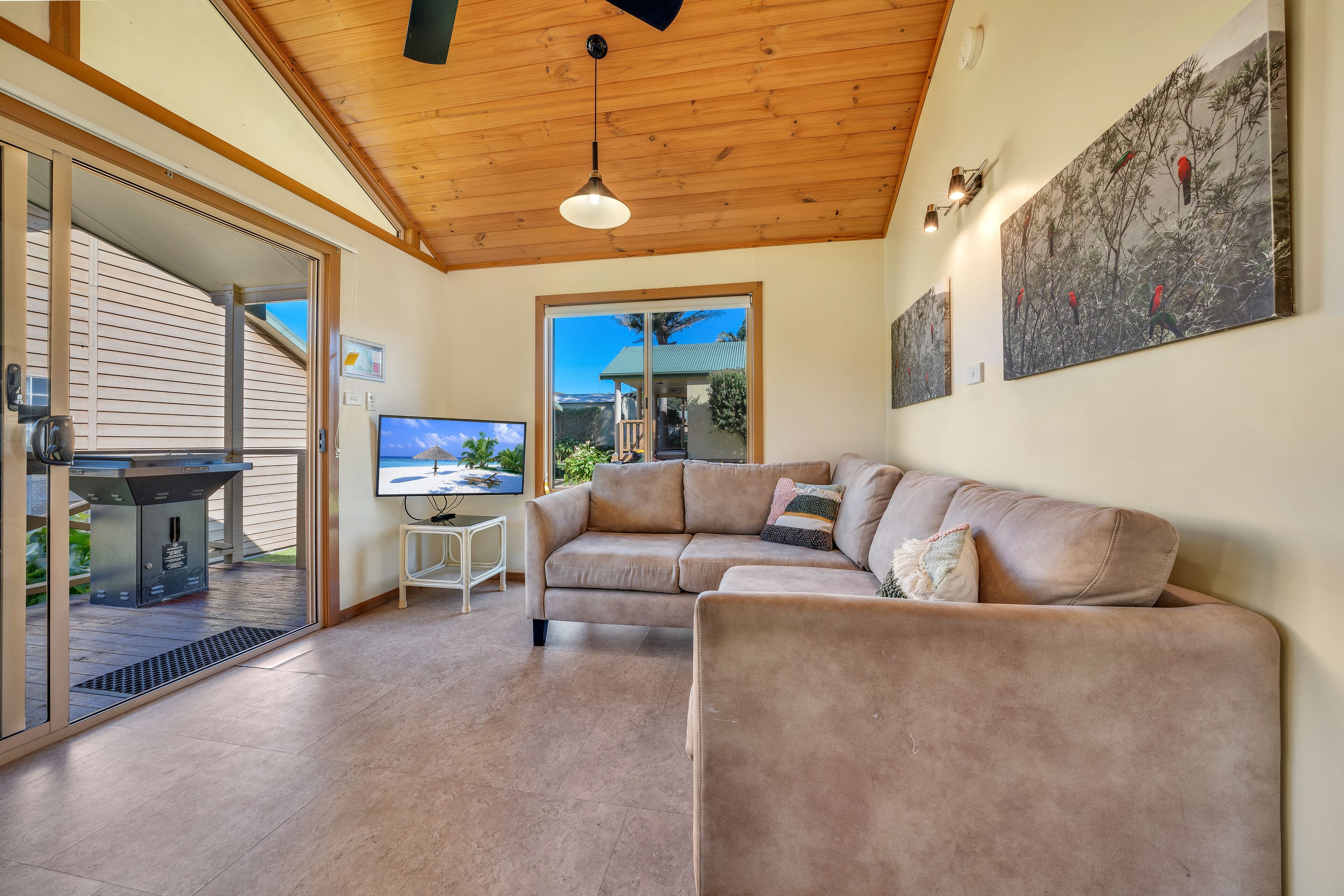 A spacious living area with a wooden ceiling, a beige sectional sofa, a coffee table, and a large window offering a view of the outdoors.