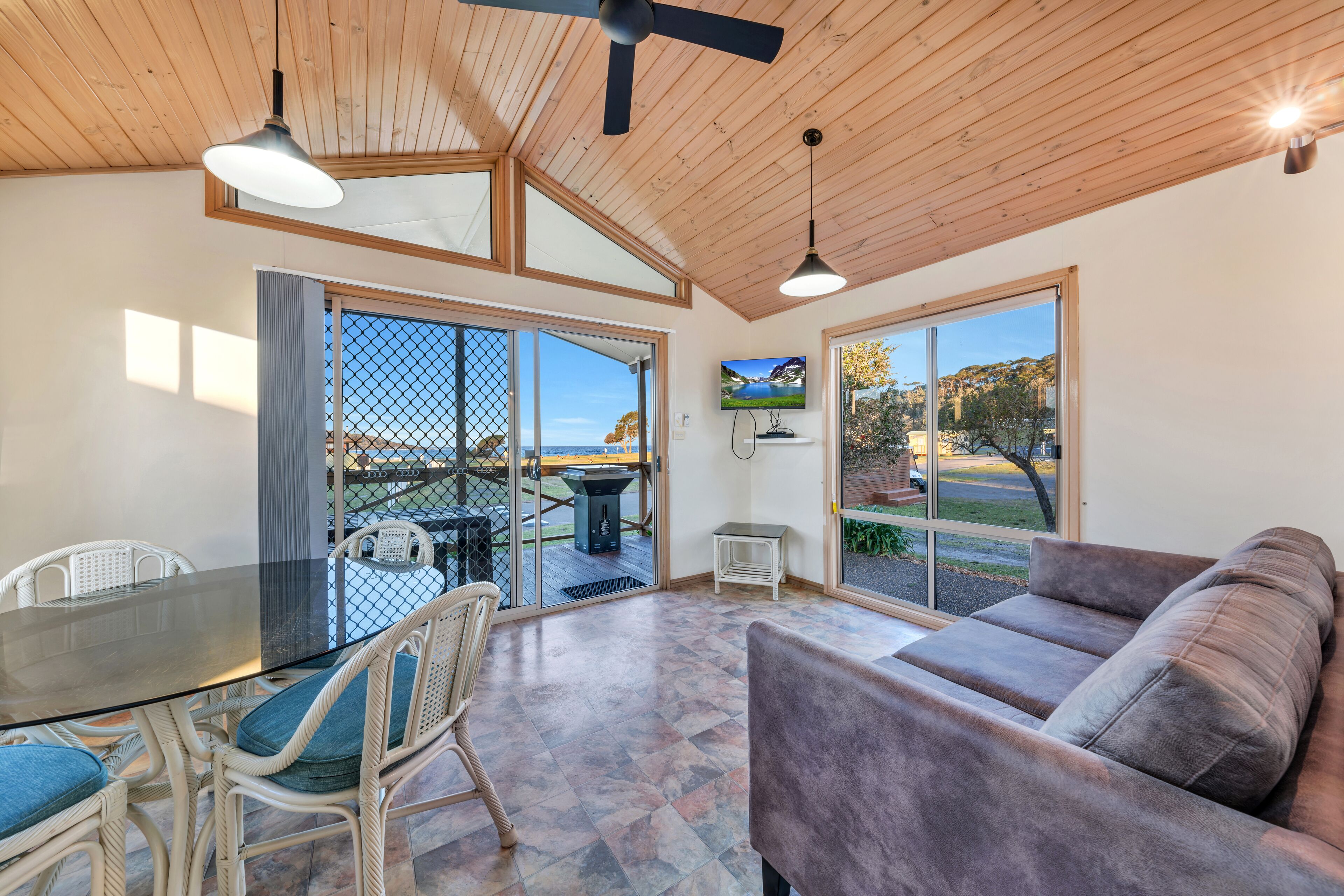 A spacious room with a glass table, wicker chairs, a cushioned sofa, and large windows offering a view of the outdoors.