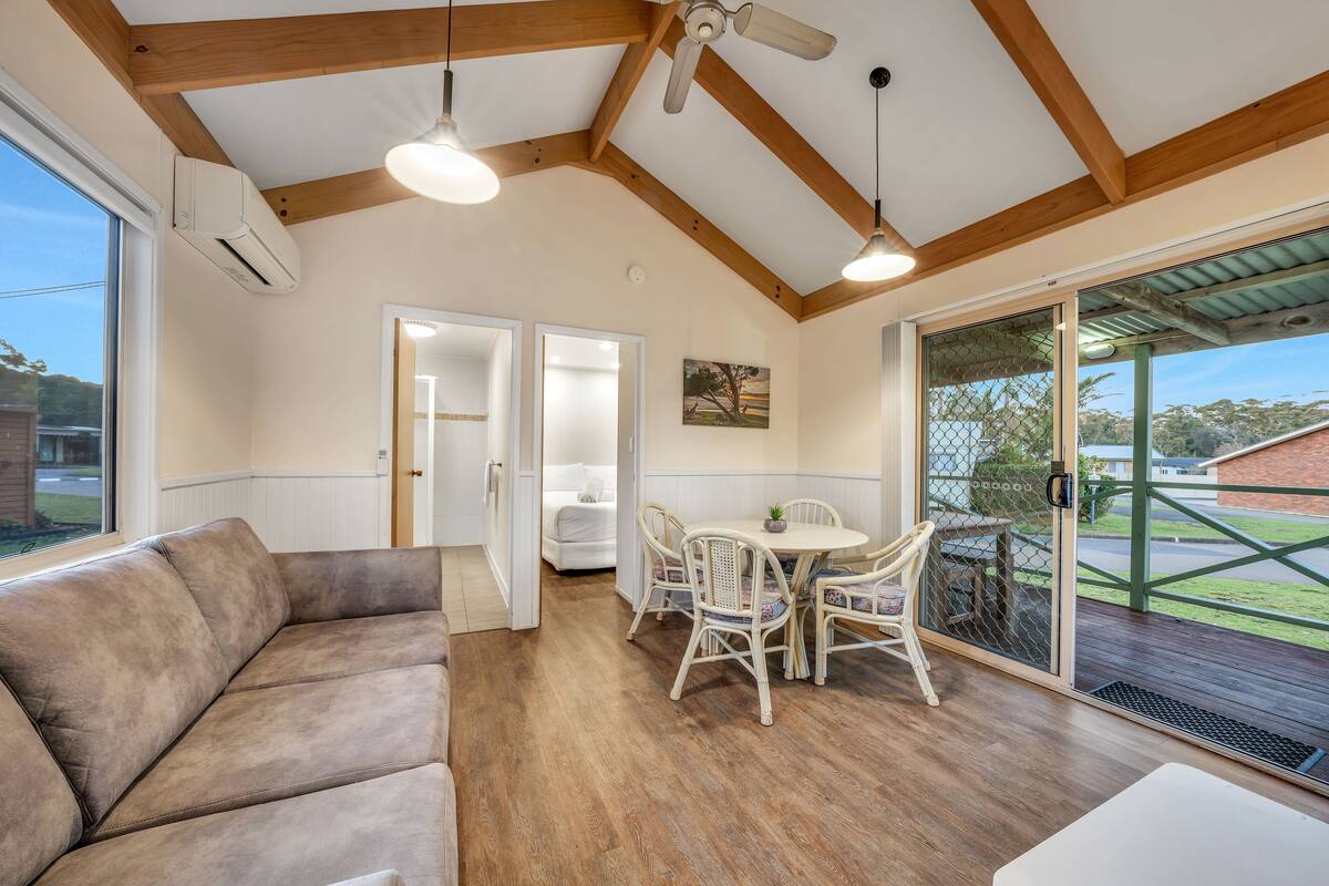 Amplia sala de estar con techo de madera, un sofá, una mesa de comedor con sillas y un ventanal corredero que da a una terraza exterior.