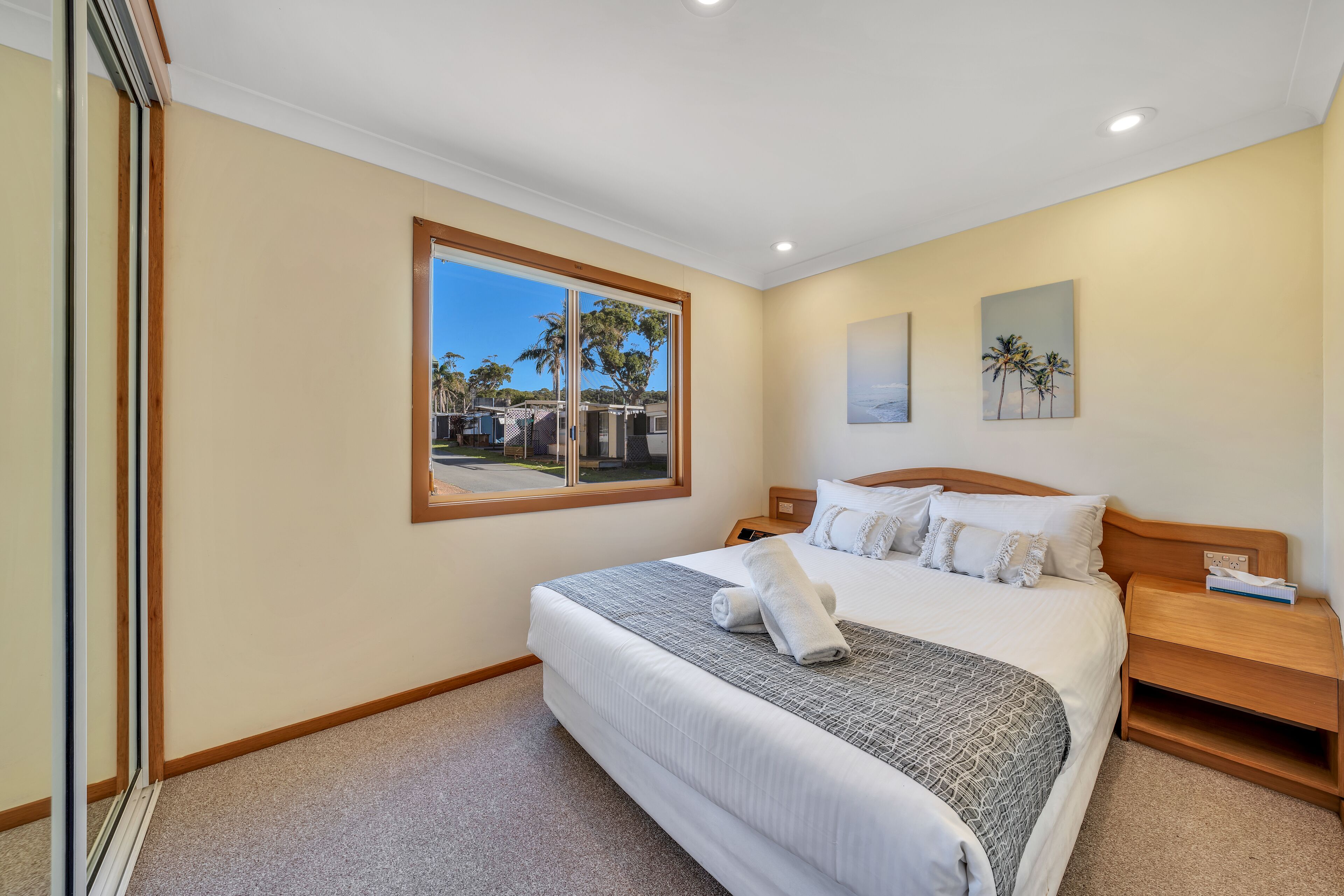 A hotel room with a large bed, a wooden headboard, a nightstand, a window with a view of palm trees, and a painting on the wall.