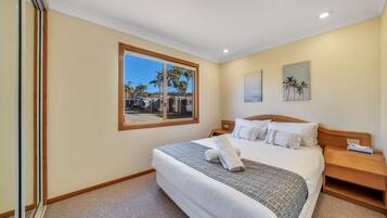 A hotel room with a large bed, a wooden headboard, a nightstand, a window with a view of palm trees, and a painting on the wall.