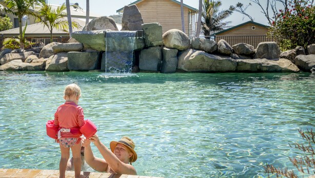 Un área de piscina con agua cristalina, rocas y un edificio de madera al fondo.