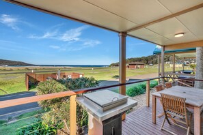 Un espacio al aire libre techado con una parrilla, una mesa de madera y sillas, con vistas a un paisaje y una playa cubiertos de césped.