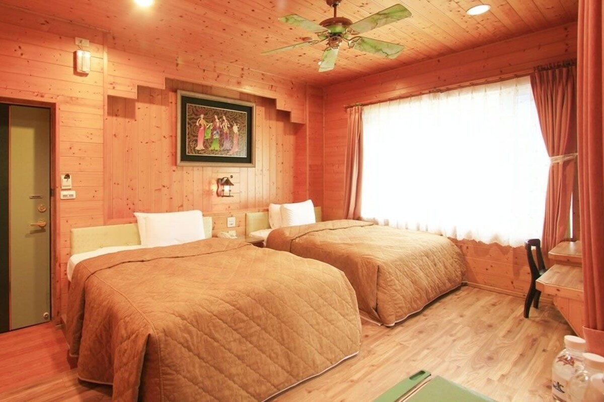 A wooden cabin room with two beds, a ceiling fan, and a large window with curtains.