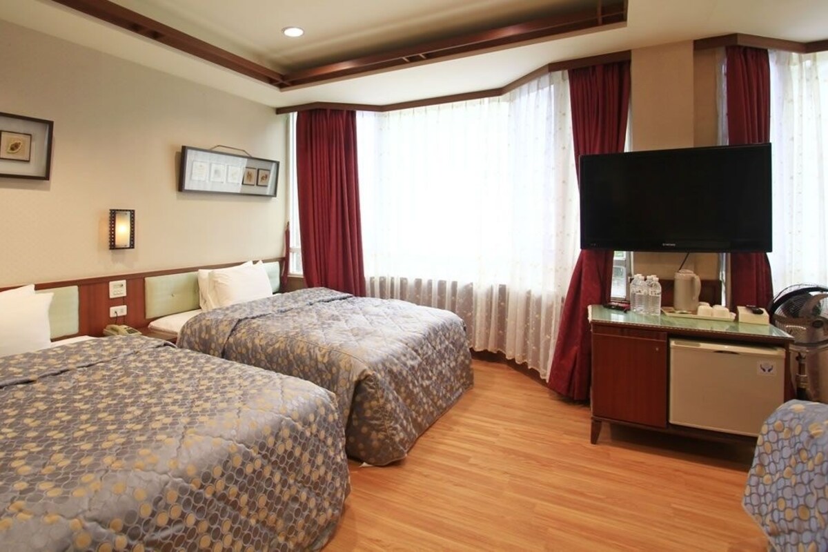 A hotel room with two beds, a TV, a desk with a fan, and a window with curtains.