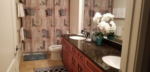 Une salle de bain avec un rideau de douche orné d’oiseaux et du mot « Espoir », des toilettes, un lavabo avec un miroir et un vase rempli de fleurs blanches.
