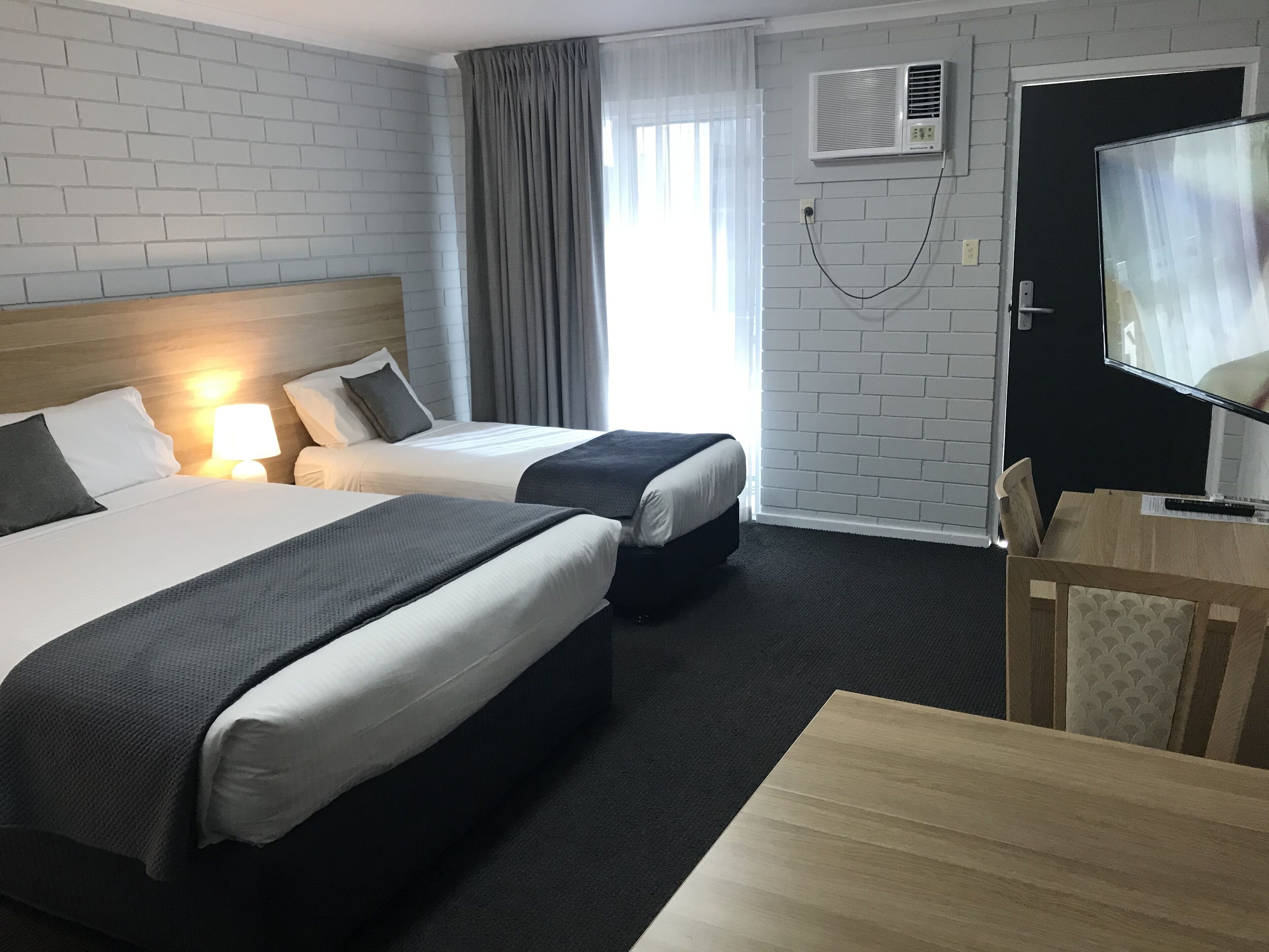 A hotel room with two beds, a desk, a chair, a TV, and an air conditioning unit.