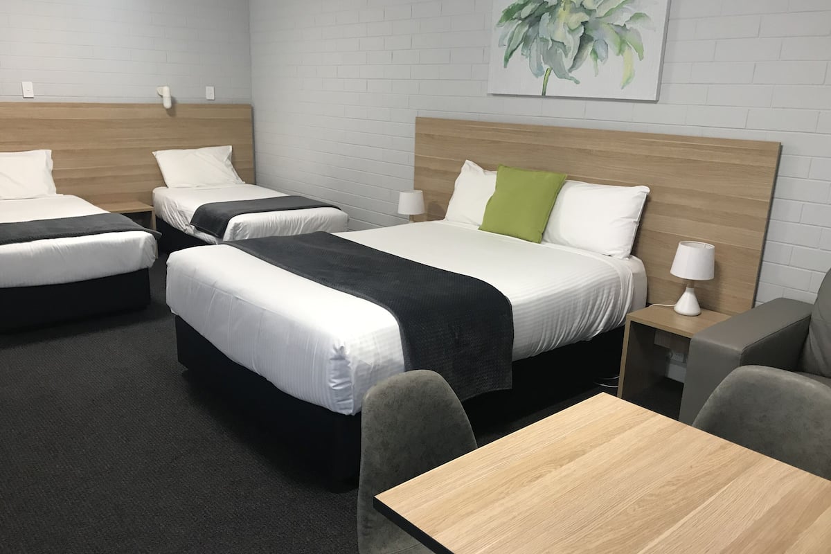 Hotellihuone, jossa on kolme sänkyä, puinen pääty, harmaa sohva, puinen pöytä ja yöpöytä, jossa on lamppu.