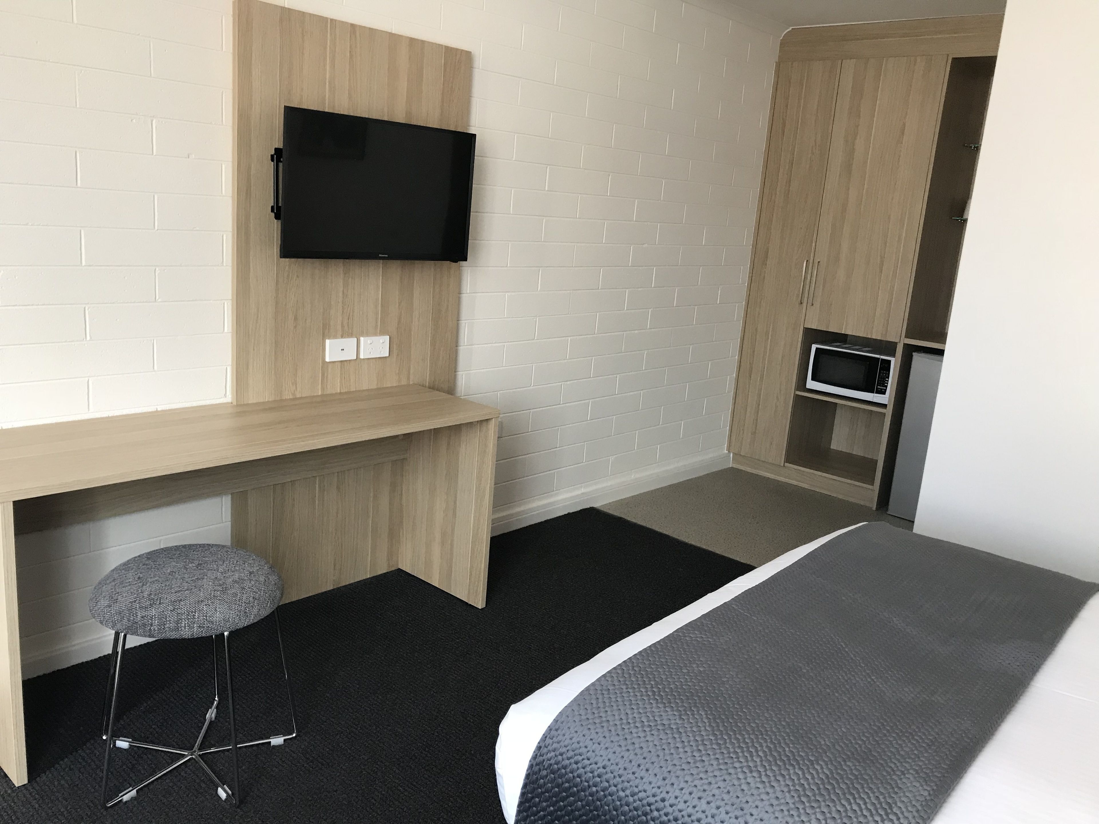 A modern hotel room with a wooden desk, a mounted flat-screen TV, a small stool, a bed with a grey headboard, and a kitchenette with a microwave and refrigerator.