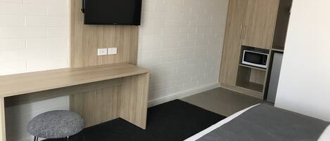 A modern hotel room with a wooden desk, a mounted flat-screen TV, a small stool, a bed with a grey headboard, and a kitchenette with a microwave and refrigerator.