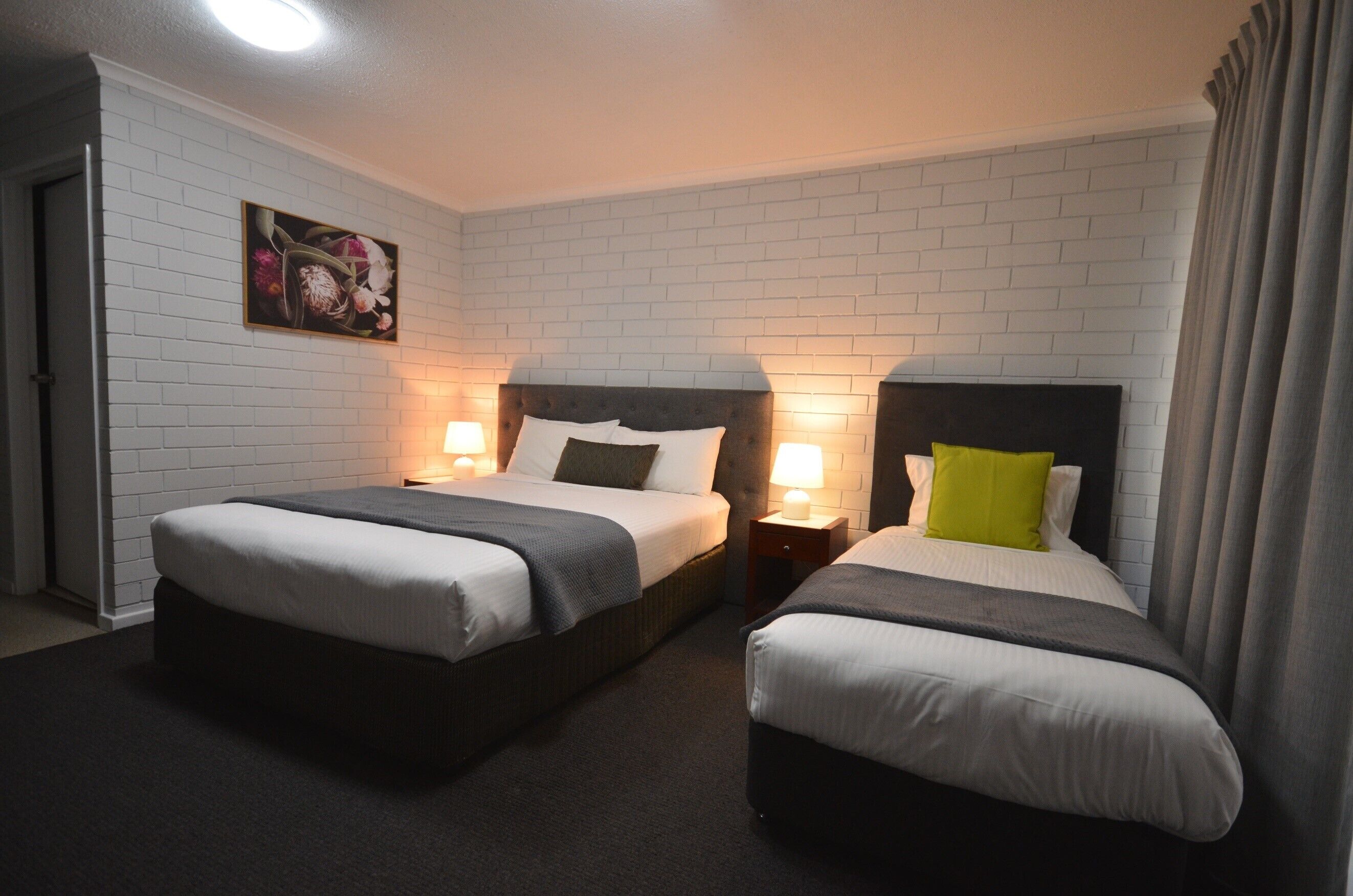 A hotel room with two beds, white brick walls, a painting on the wall, and a door to the left.