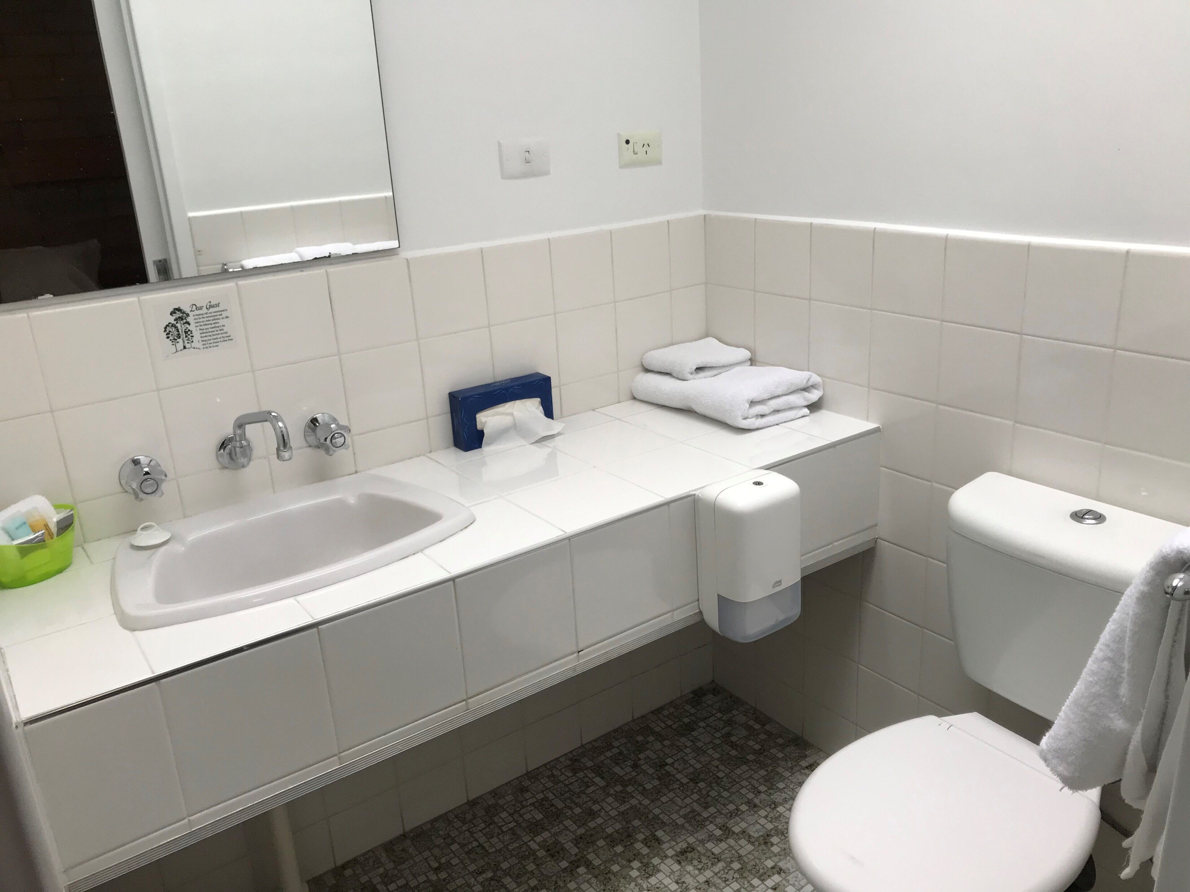 A bathroom with a white sink, a toilet, and a mirror. There is a towel dispenser and a tissue box on the counter.