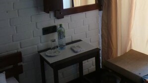 Habitación de hotel con pared de ladrillo blanco, espejo con marco de madera, una mesita con una botella de agua, una cama y una ventana con cortinas color beige.