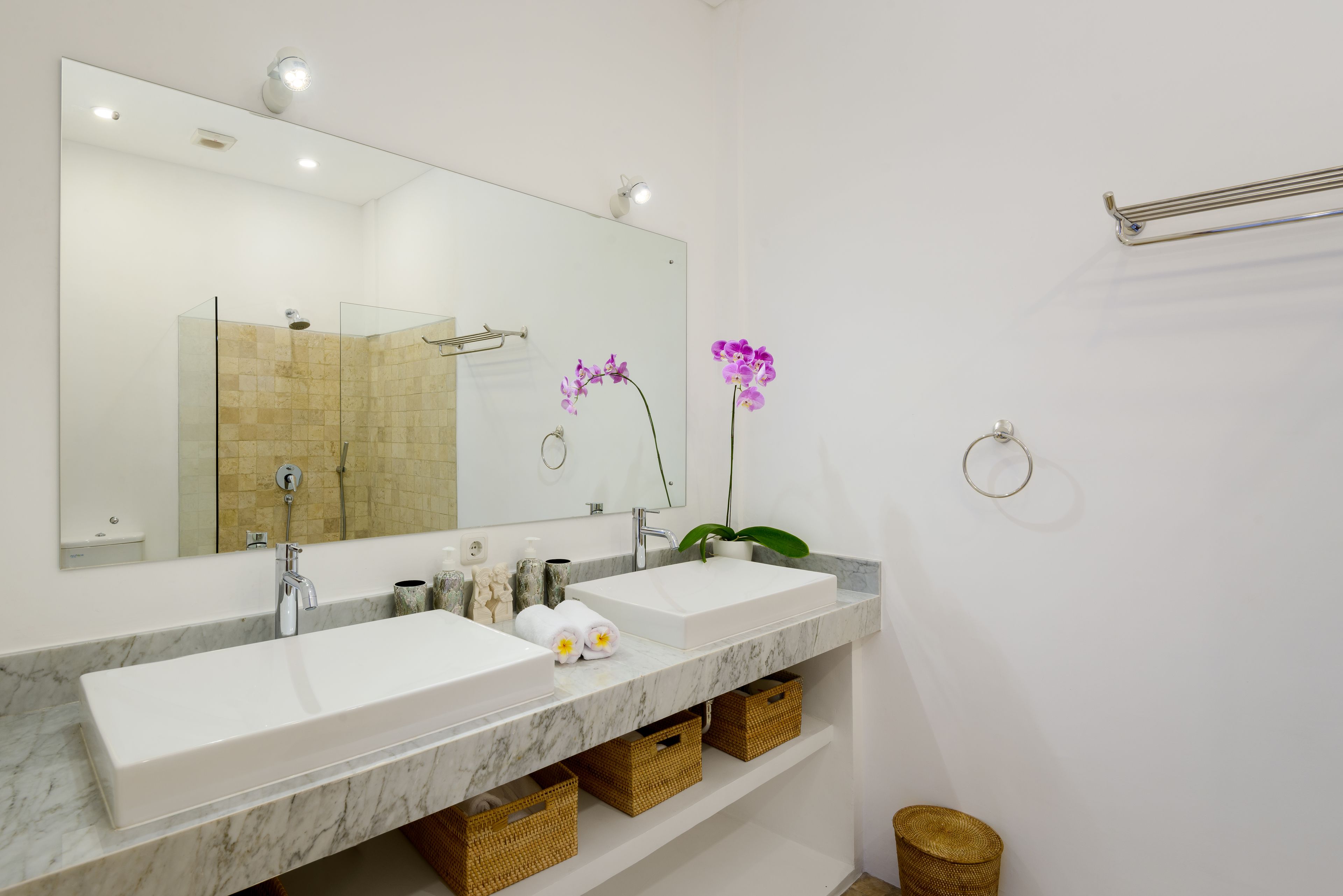 Une salle de bain avec deux lavabos, un grand miroir et une douche. On trouve des paniers en osier sous les lavabos et un petit panier en osier sur le sol.