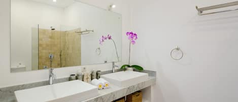 Une salle de bain avec deux lavabos, un grand miroir et une douche. On trouve des paniers en osier sous les lavabos et un petit panier en osier sur le sol.