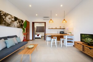 A modern living room with a sofa, coffee table, and a TV. There is a kitchen area with a dining table and bar stools.