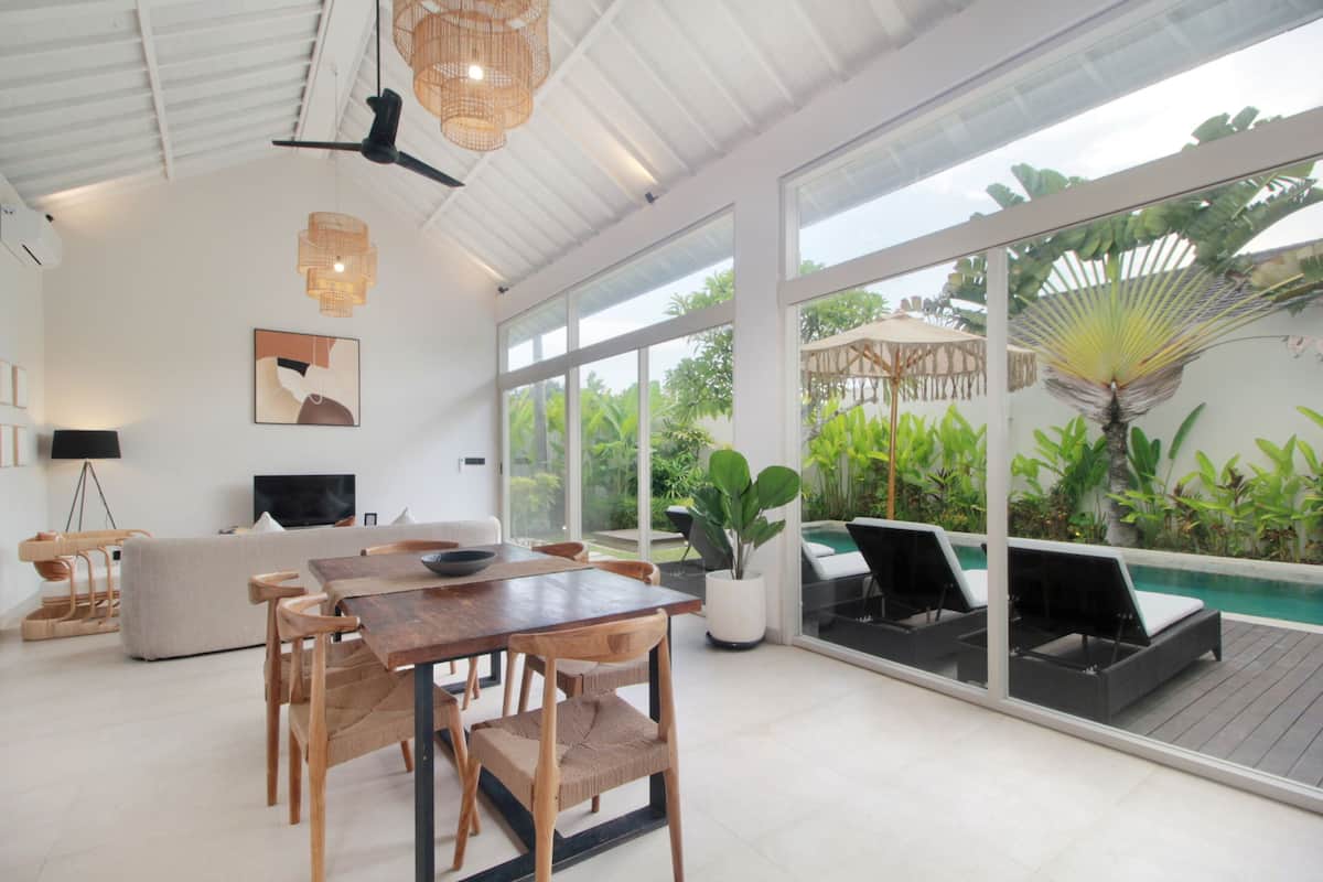 A spacious living area with a dining table, chairs, a sofa, and a TV. There is a large window offering a view of a pool and lush greenery.