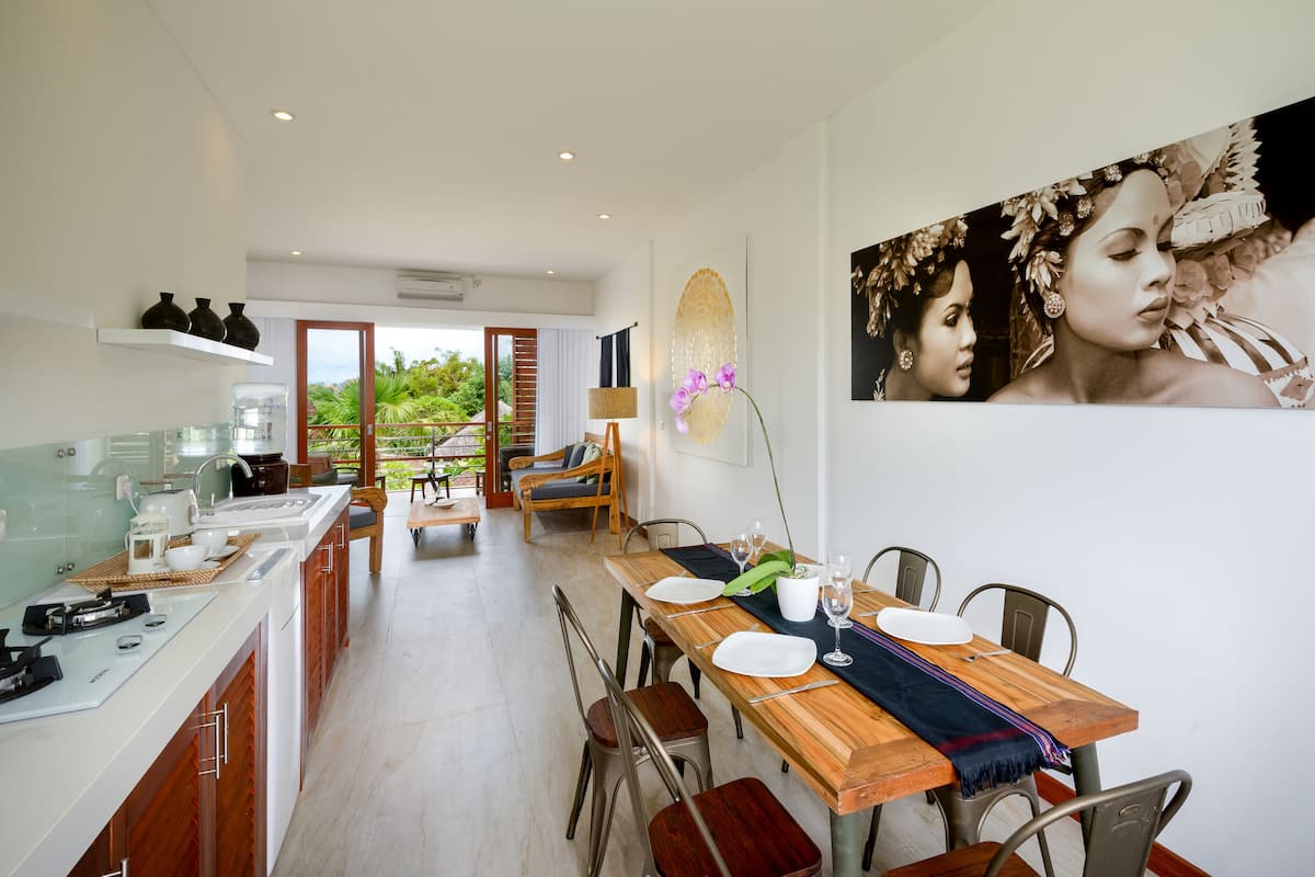 A modern kitchen with a dining area, wooden table, chairs, and a view of greenery outside.