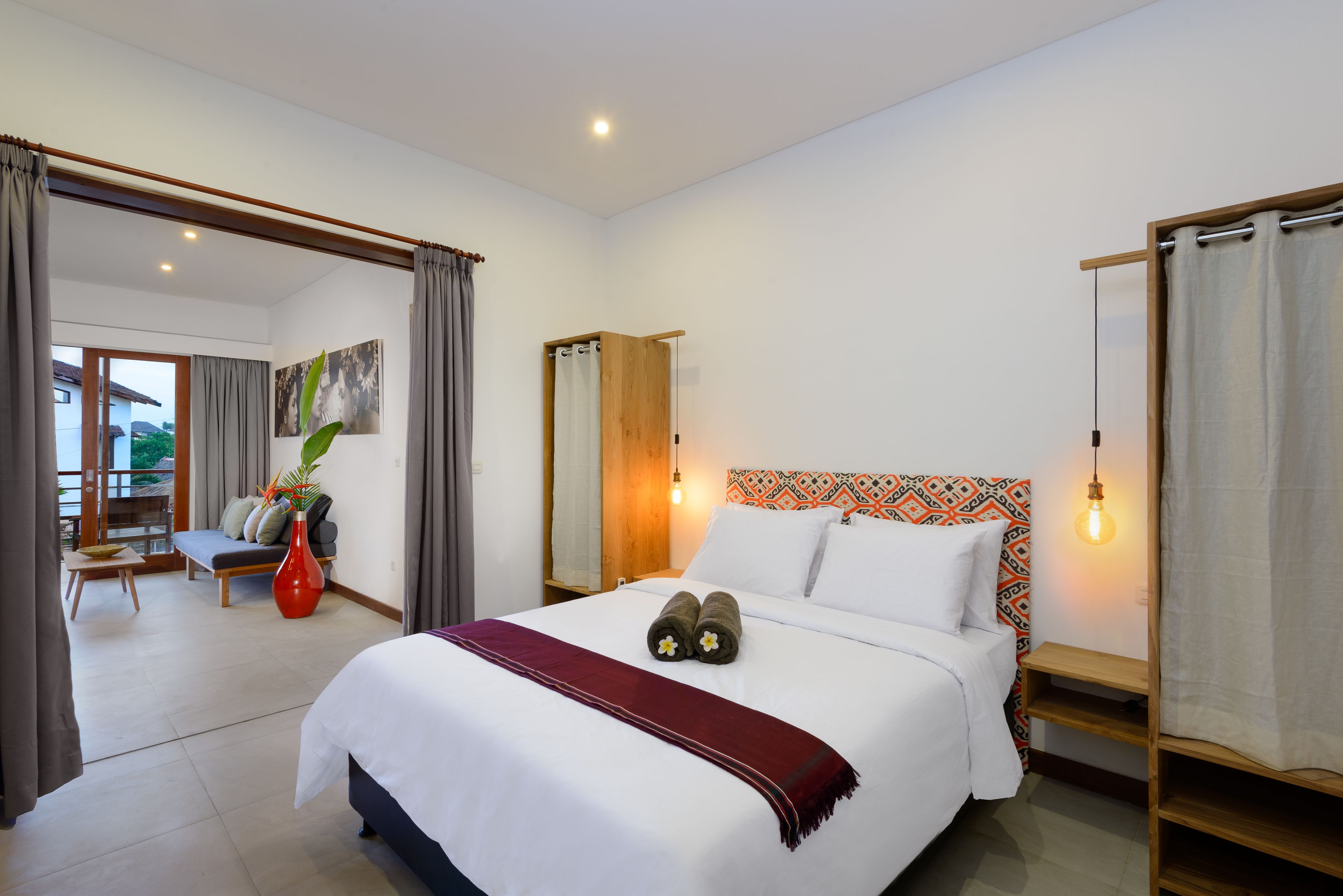 A hotel room with a bed, a bench, a red vase, and a view of the outside.