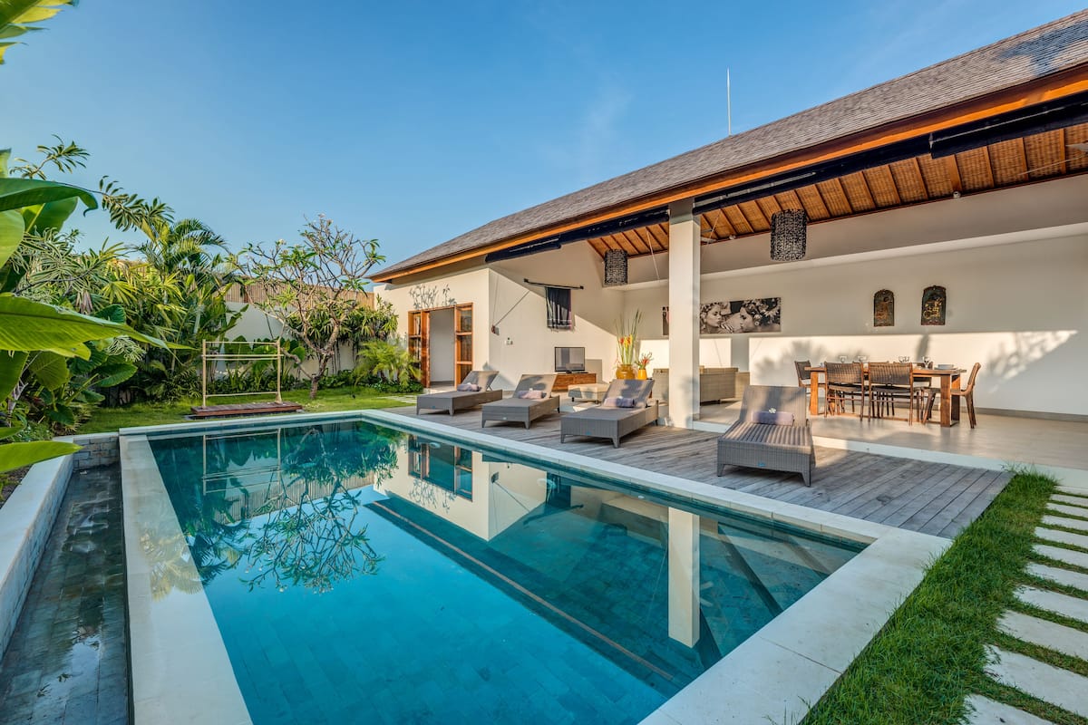 A modern outdoor pool area with lounge chairs and a wooden deck, surrounded by lush greenery.